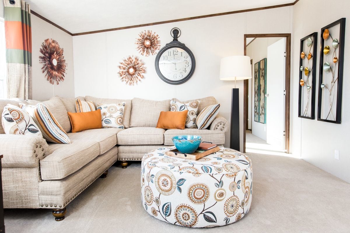 Cozy living room with a beige sectional sofa adorned with orange and patterned pillows. A round, floral ottoman with a blue bowl sits in front. The walls display a large clock and sunburst decor, alongside vertical art with metallic accents. The setting is inviting and stylish.