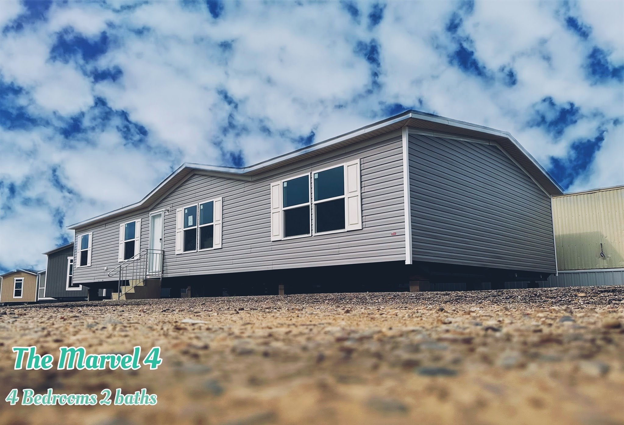 Single-story modular home with grey siding under a blue sky with clouds. A sign in the foreground reads "The Marvel 4, 4 Bedrooms 2 baths."