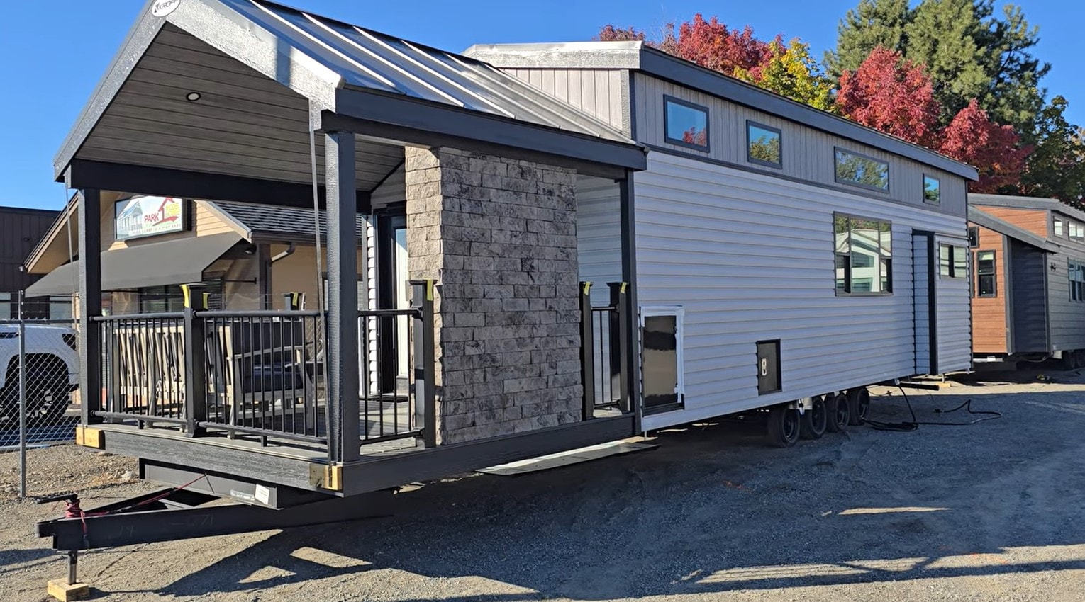 A modern tiny house on wheels with a stone-accented porch and metal roof, set against autumn trees. The scene conveys a cozy, innovative lifestyle.