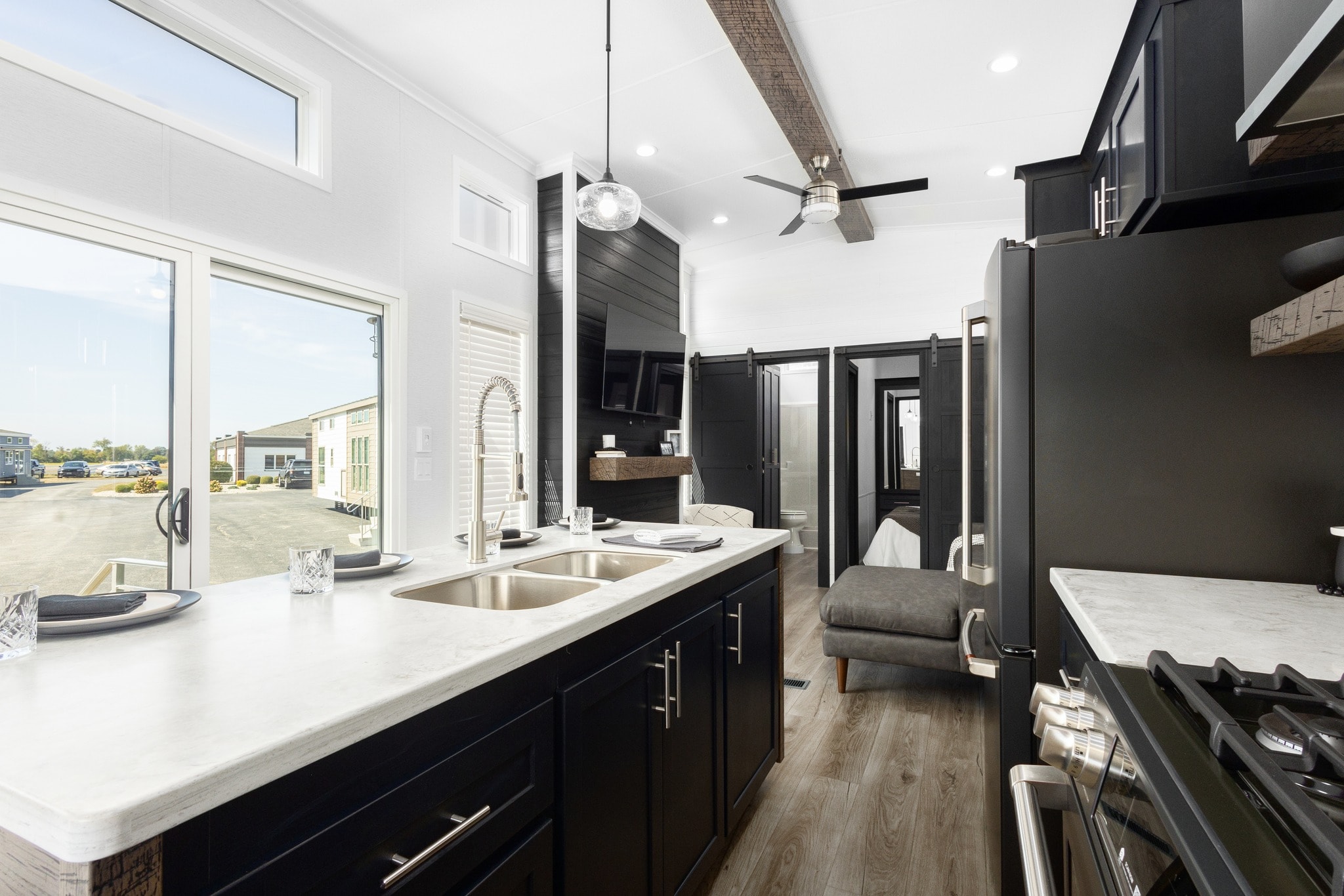 Modern kitchen with sleek black cabinets, white countertops, and stainless steel appliances. A large window offers natural light, creating an airy feel.