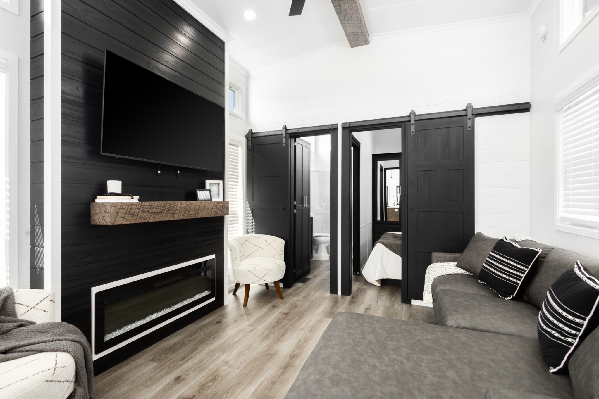 Modern living room with black paneled walls, a wall-mounted TV, and a linear fireplace. Gray sofa, patterned cushions, and barn doors add a cozy touch.