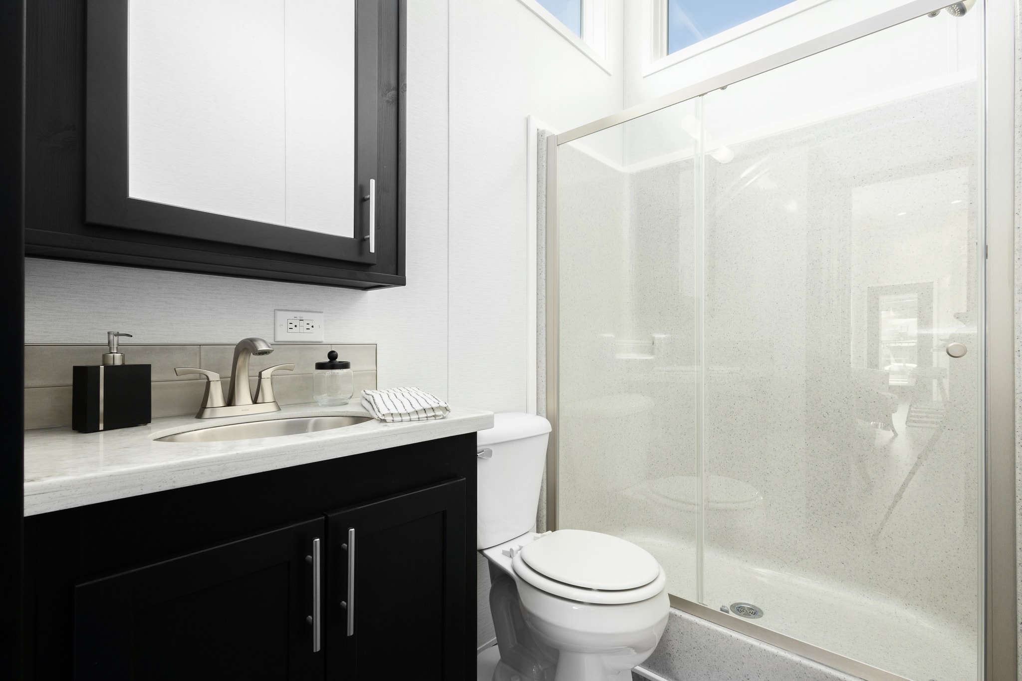 Modern bathroom with a sleek design features a black vanity with a silver faucet, white countertop, toilet, and clear glass shower door under a bright skylight.