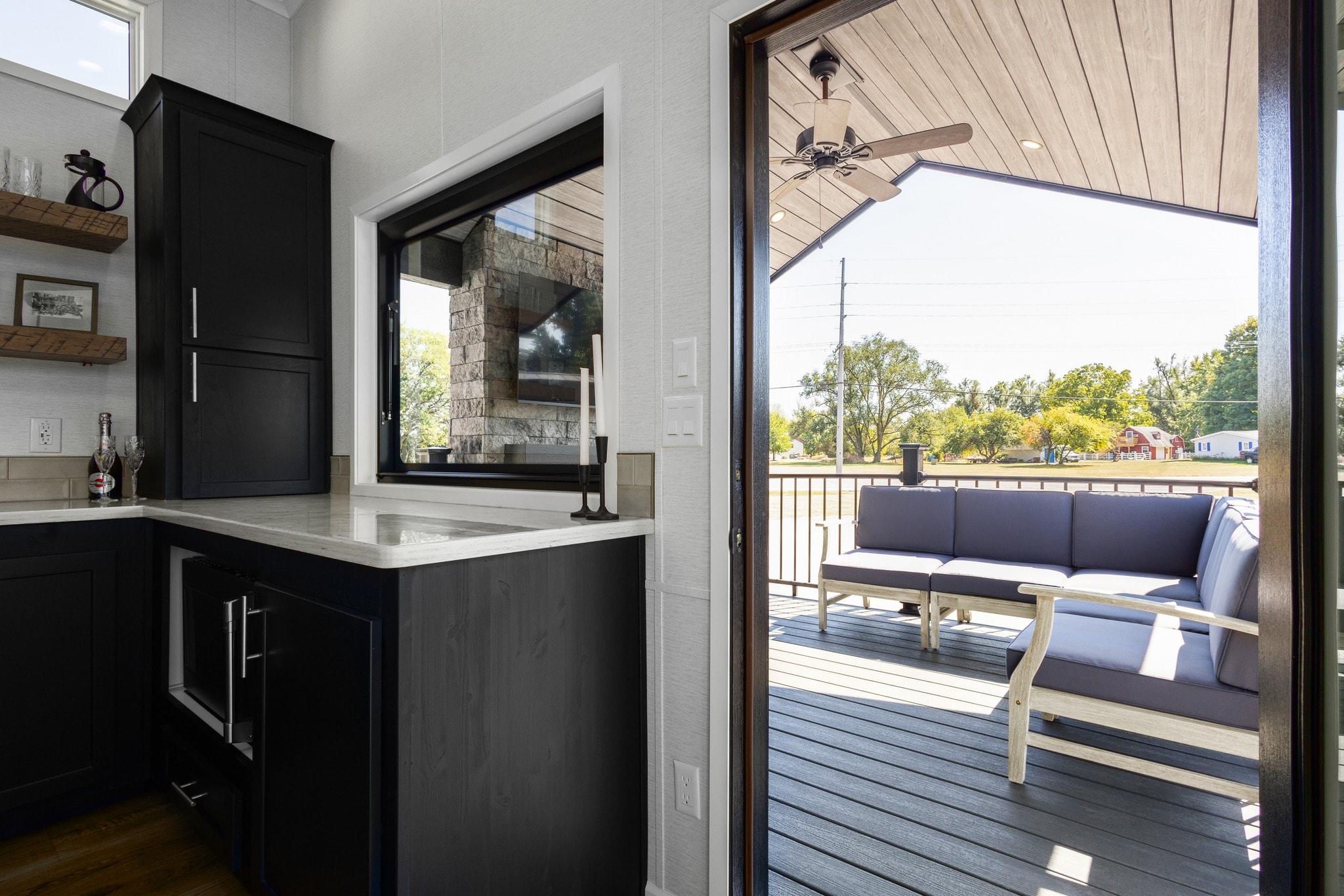 Modern kitchen with dark cabinetry and open shelves on the left. Through a door, a cozy patio with blue seating and wooden floors is visible outside.