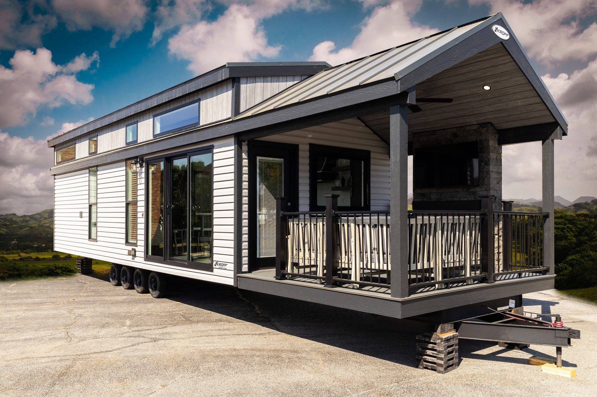 Modern tiny house on wheels with white siding and dark trim, featuring large windows and a covered porch. Set against a scenic mountain background.