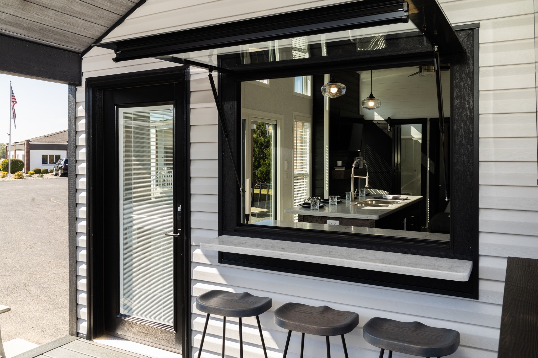 Outdoor bar setup with three stools beneath a black-framed window and a glass door. Modern kitchen visible inside. Bright, inviting atmosphere.