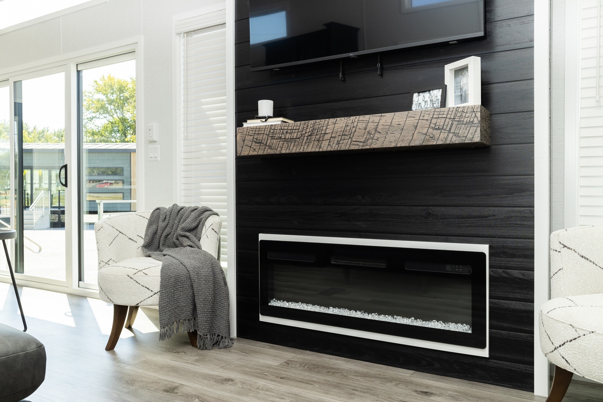 Modern living room with a sleek black fireplace under a wall-mounted TV. Light wood flooring, cozy armchair with a gray blanket, and large windows.