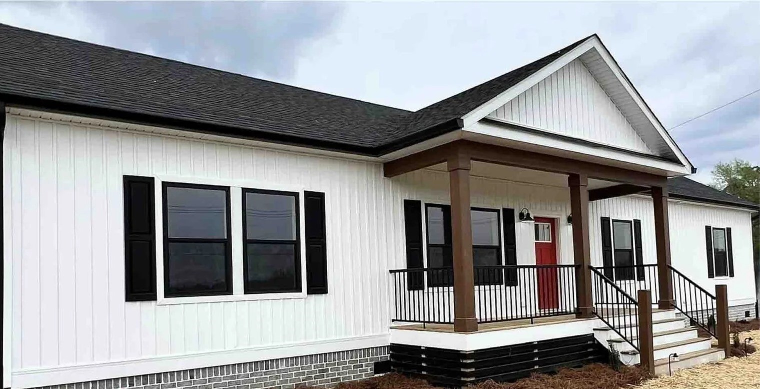 A modern white house features black trim and shutters, a bright red door, and a welcoming porch with brown columns. The sky is overcast.