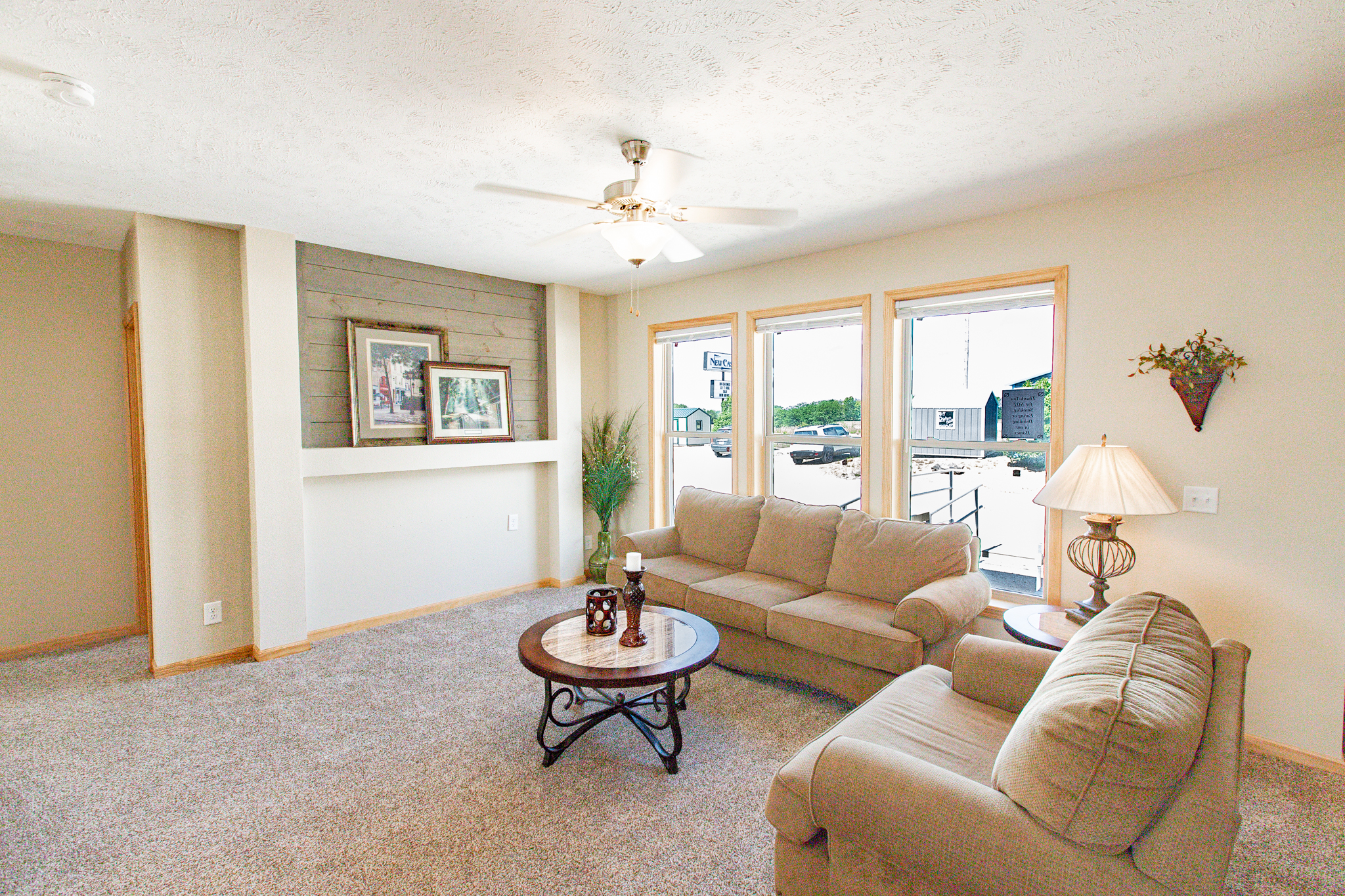 Bright living room with beige walls and carpet, featuring a cozy sofa, two tables with lamps, wall art, and large windows for natural light.