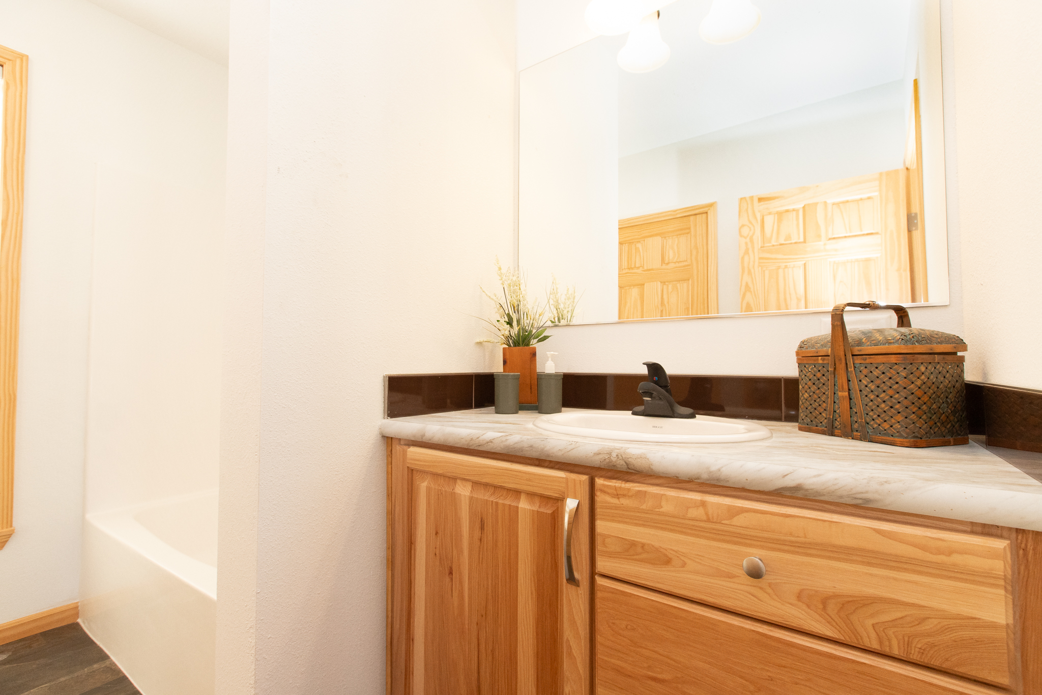 Bright bathroom with light wood cabinets, a marble countertop with a sink, potted plant, and wicker basket. Soft lighting and warm tones create a cozy feel.