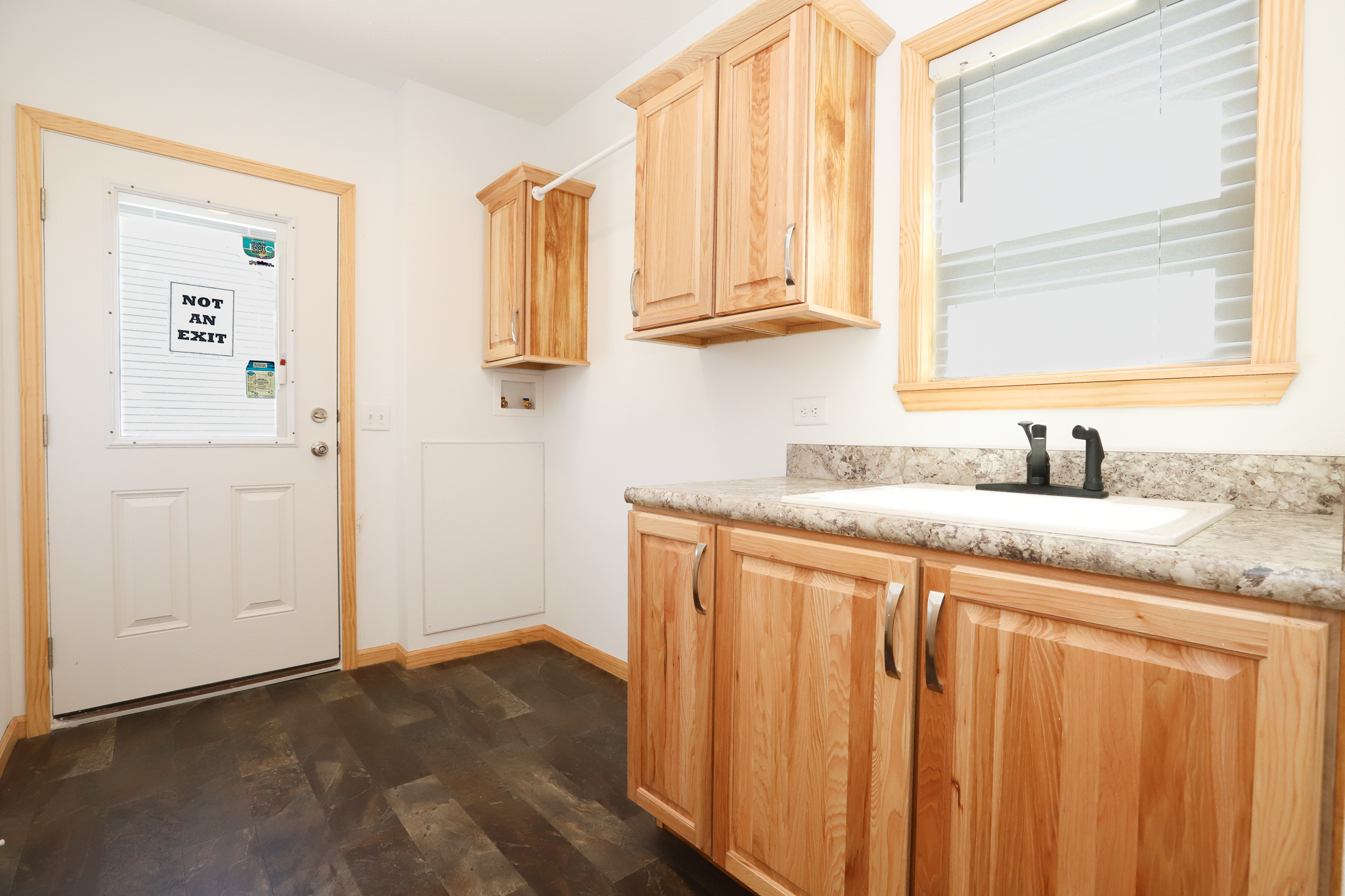 Bright utility room with wooden cabinets, a granite countertop with a sink, and dark tile flooring. Door labeled "Not an Exit" and a window with blinds.