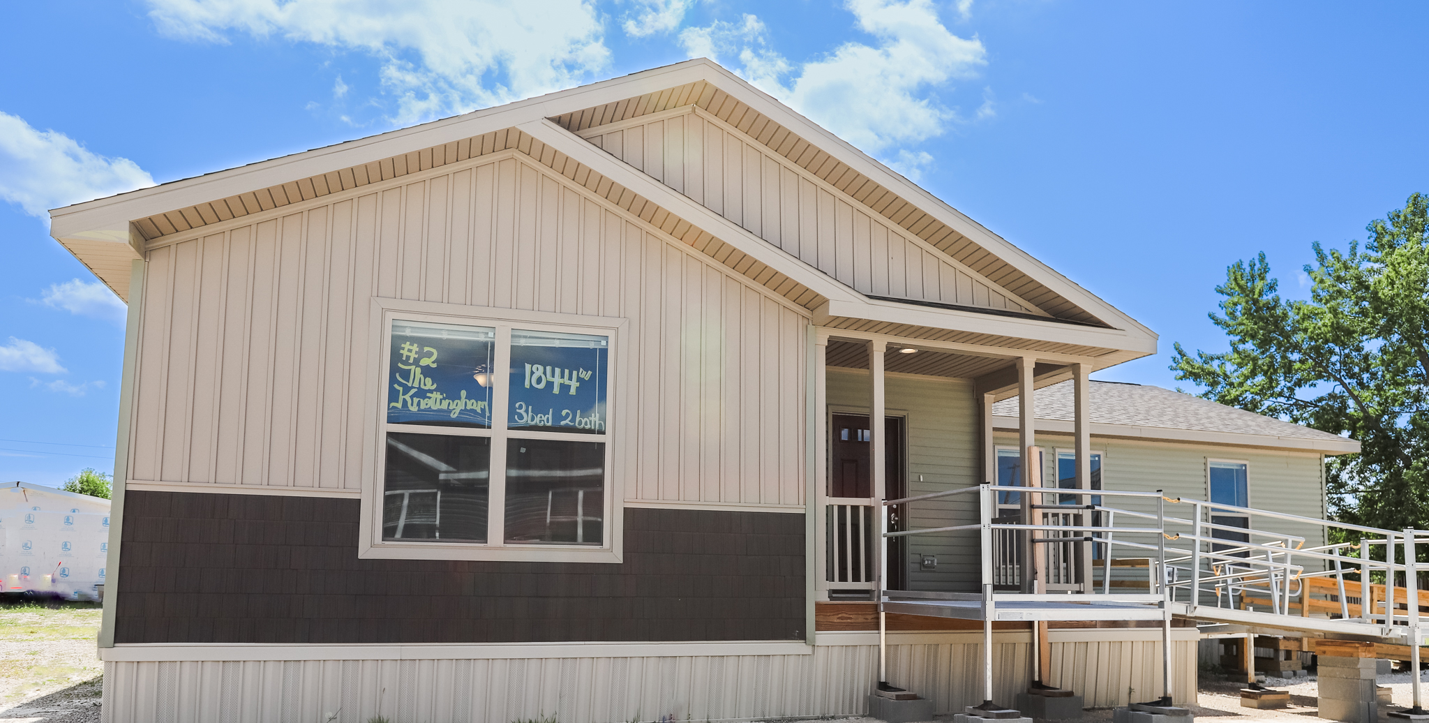 A modern single-story house with beige siding, a wooden ramp, and a sloped roof under a bright blue sky. Large windows have handwritten details.
