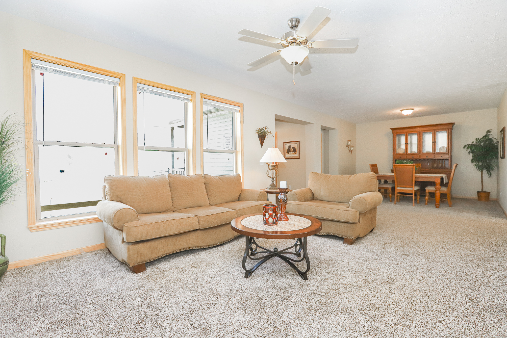 Living room with beige sofas, round coffee table, and large windows. Ceiling fan above and dining area in the background, creating a cozy, airy feel.