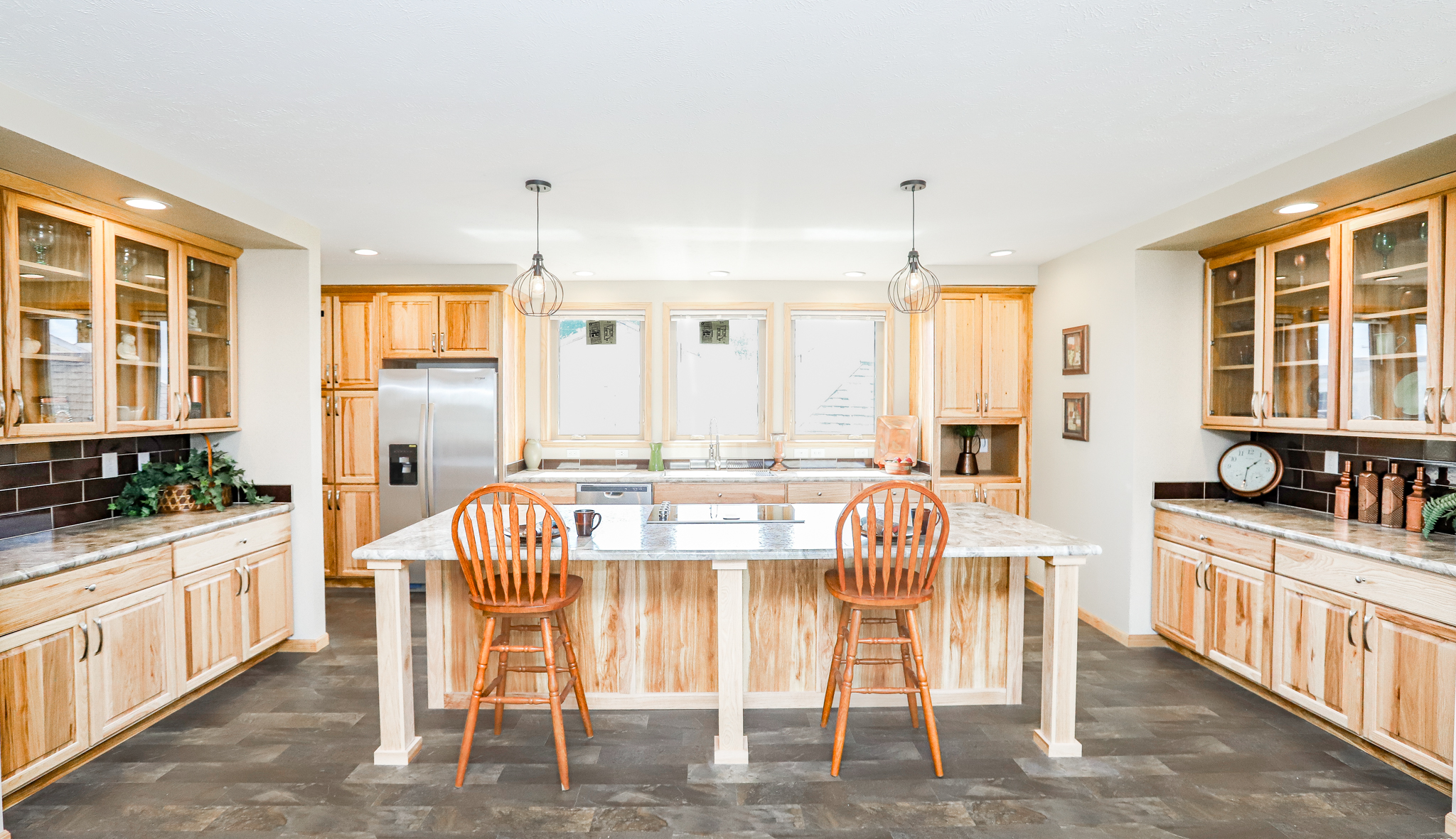 Bright, modern kitchen with wooden cabinets, marble countertops, and two wooden stools at the island. Pendant lights hang overhead. The space feels warm and inviting.