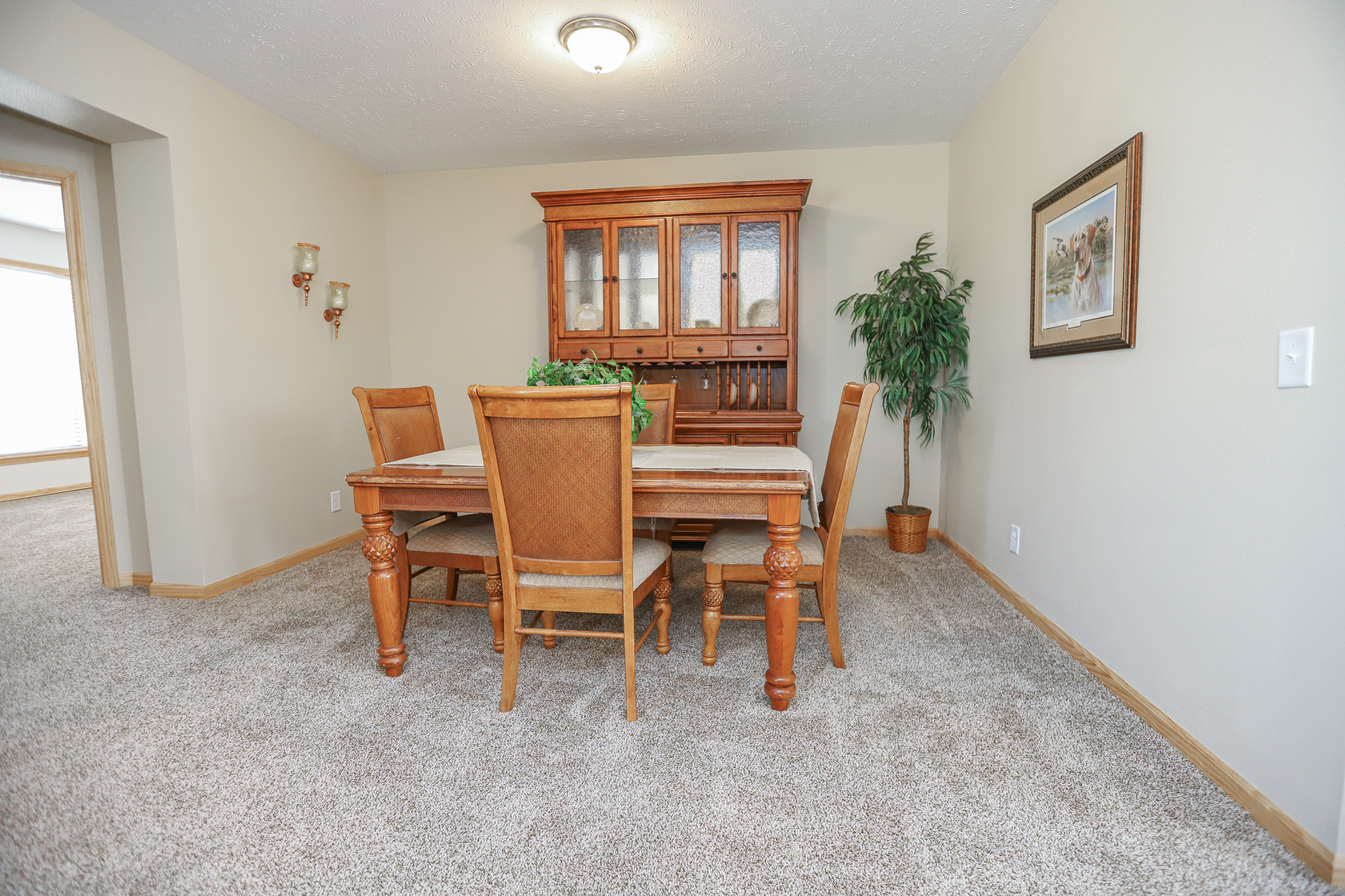 A cozy dining room with a wooden table and four chairs, a potted plant, and a wooden cabinet. The room has warm lighting and carpeted flooring.