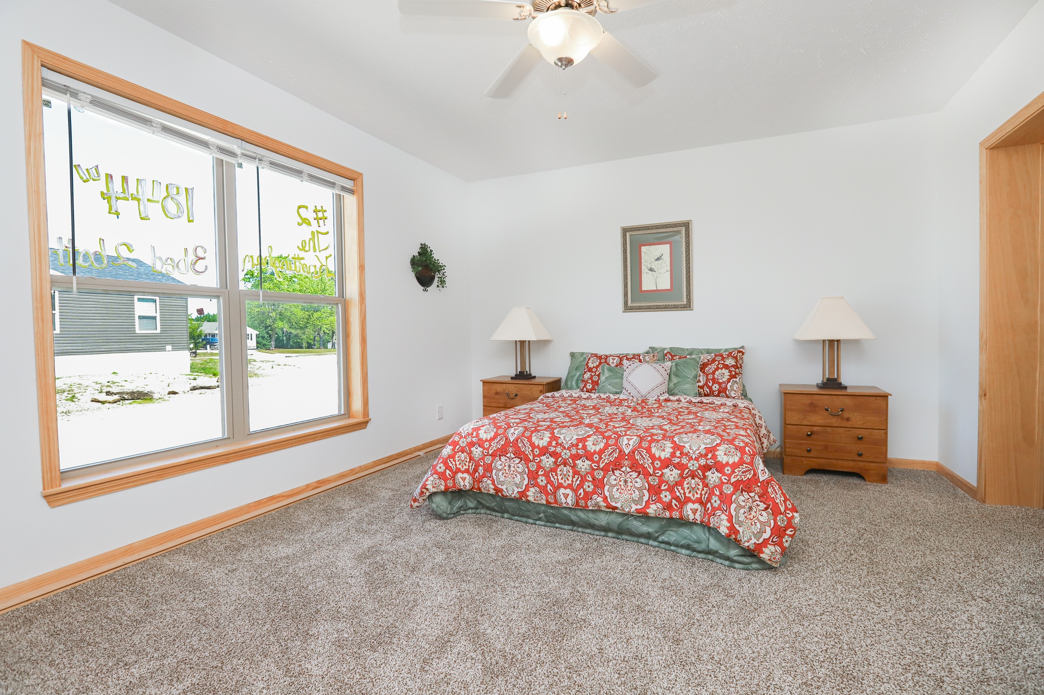 Bedroom with a white ceiling and light gray walls, featuring a bed with a red floral comforter between two wooden nightstands and lamps. Large window on the left.