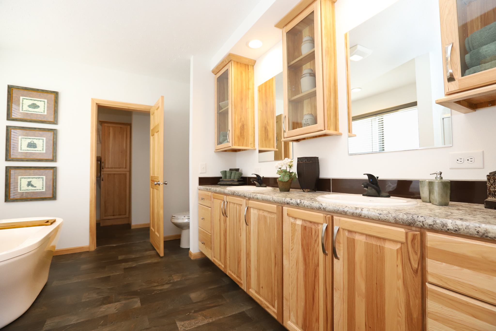 Spacious bathroom with natural wood cabinets and dark countertops. A white bathtub is visible on the left. Soft lighting and wall art create a cozy feel.