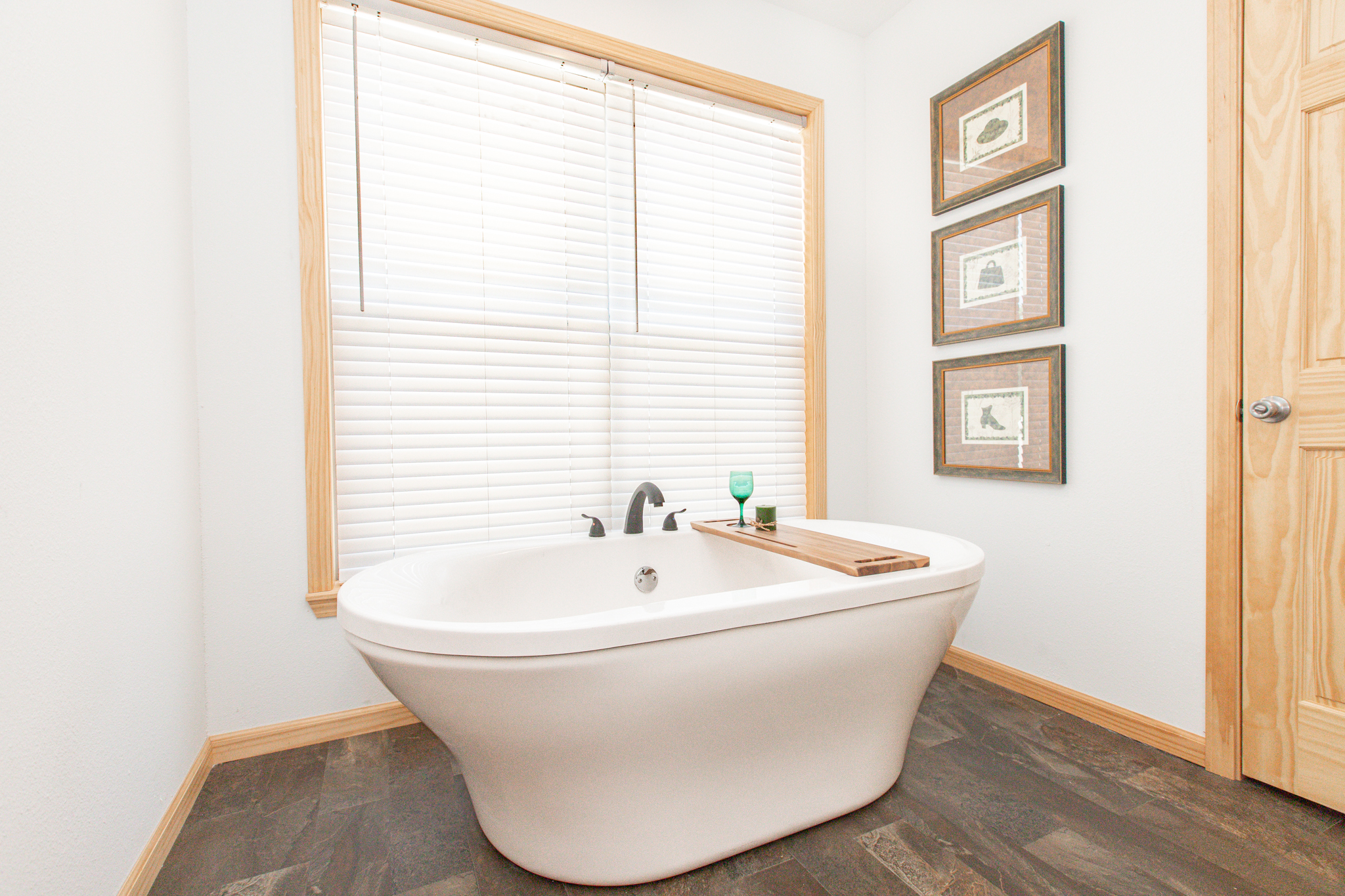 A bright bathroom with a standalone white bathtub features a wooden bath tray holding a green glass and candle. Three framed pictures hang on the wall, and a large window with blinds adds natural light, creating a serene atmosphere.