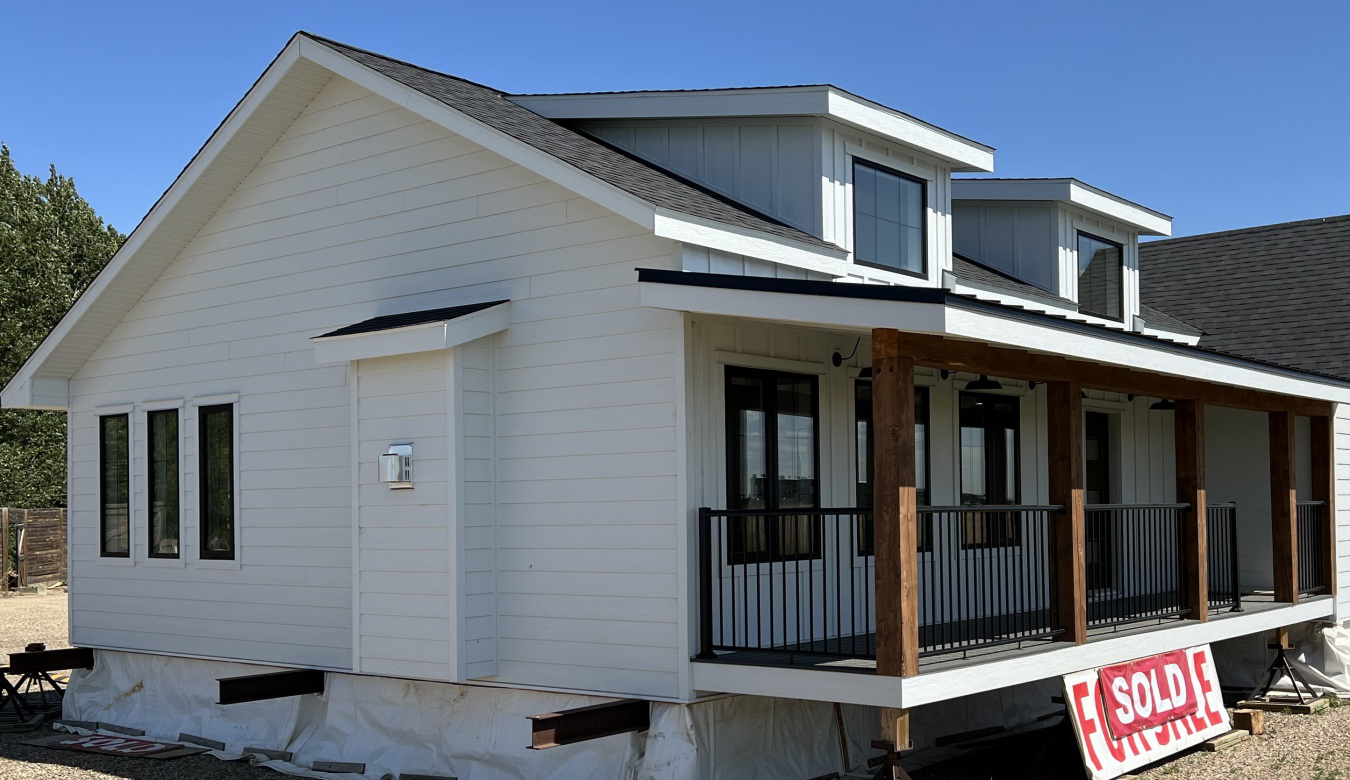 A small, modern white house with a black roof and covered porch, featuring large windows. A "Sold" sign is visible, indicating a recent sale.