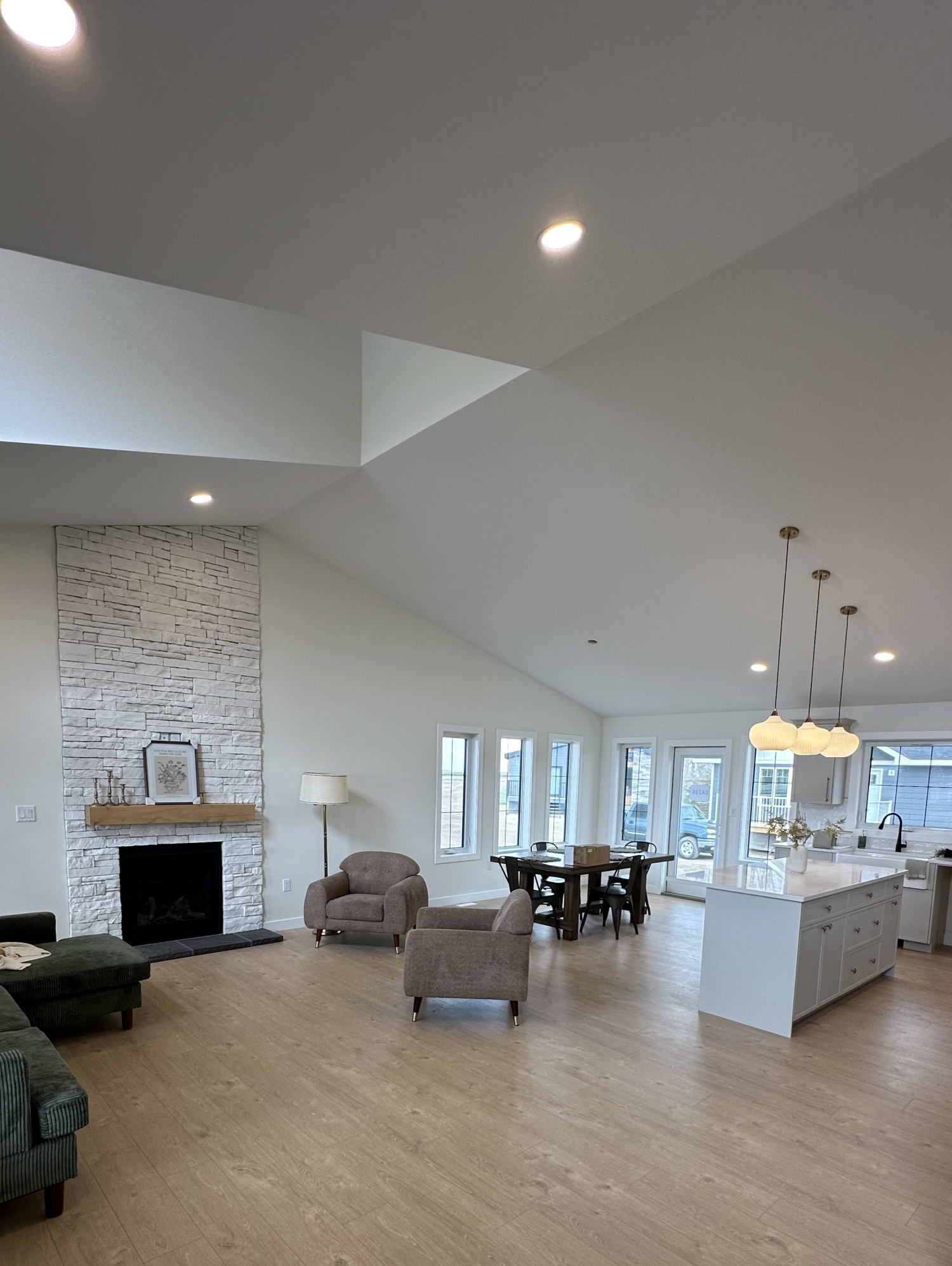 Spacious, modern living room with vaulted ceiling, stone fireplace, cozy chairs, dining table, and a kitchen island with pendant lights. Bright and inviting atmosphere.