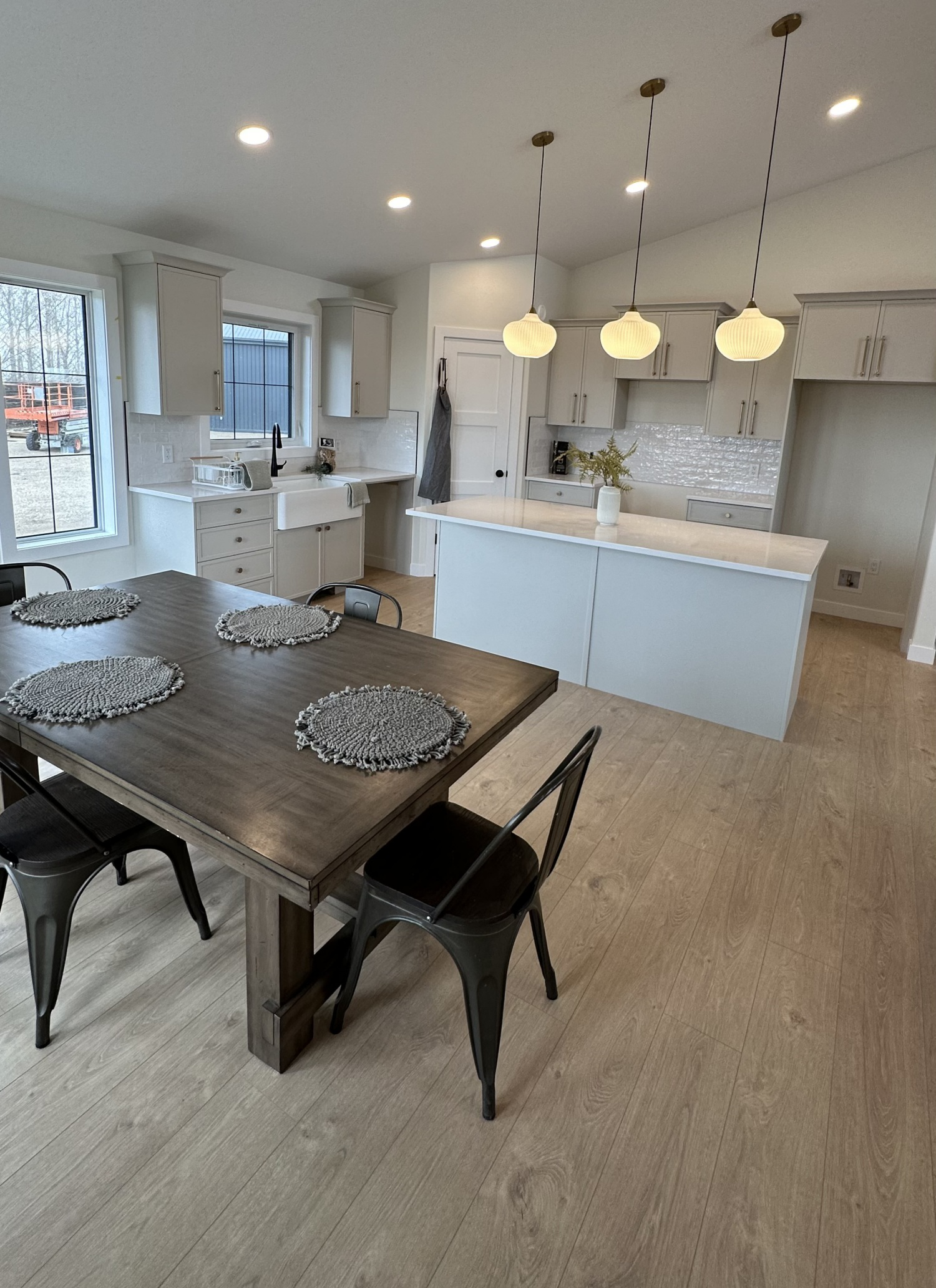 Modern kitchen with a light, airy feel. Features a wooden dining table with woven placemats, metal chairs, white cabinets, and pendant lights above an island.