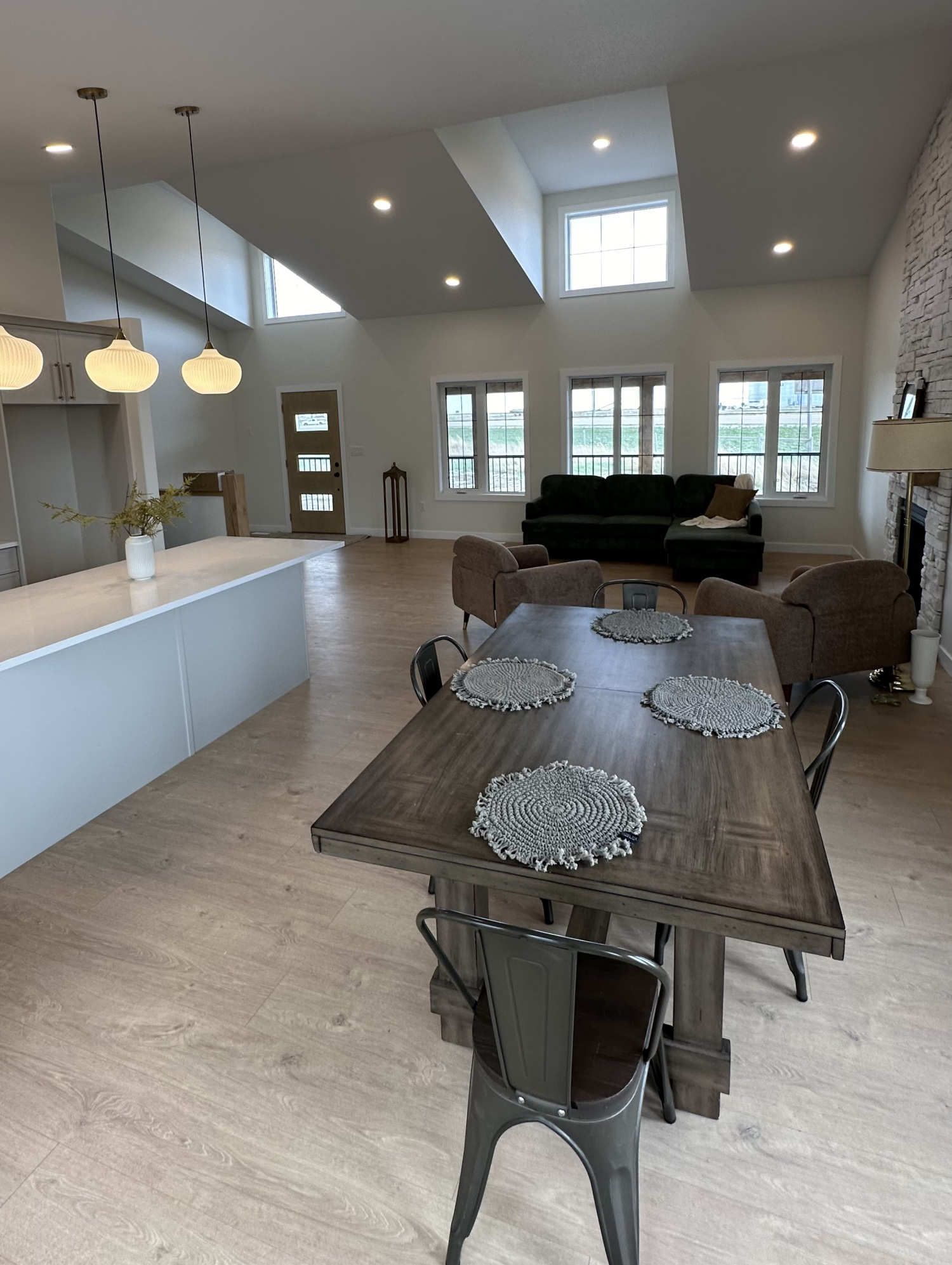 Spacious modern living area with high ceilings, large windows, and soft lighting. Features a wooden dining table, chairs, cozy sofas, and a fireplace.