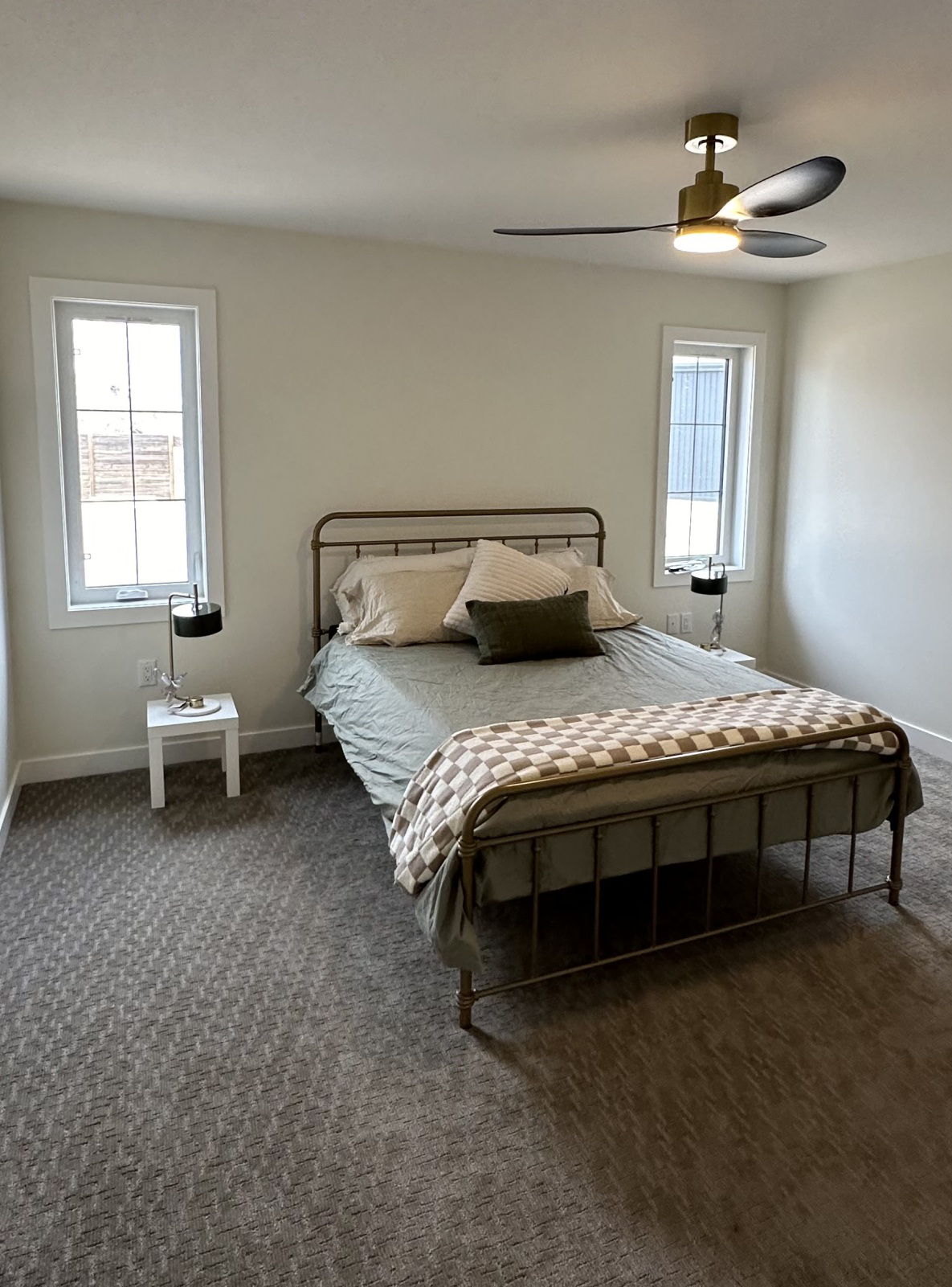 Minimalist bedroom with a metal bedframe, green bedding, and a checkered throw. Two windows with side tables, dark lamps, and a ceiling fan. Peaceful tone.