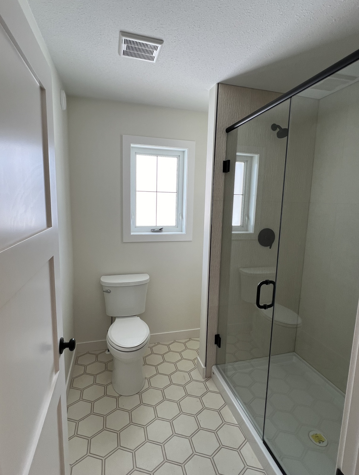 A small bathroom with neutral tones features a window, toilet, and glass shower enclosure. Hexagonal tile flooring adds a modern touch.