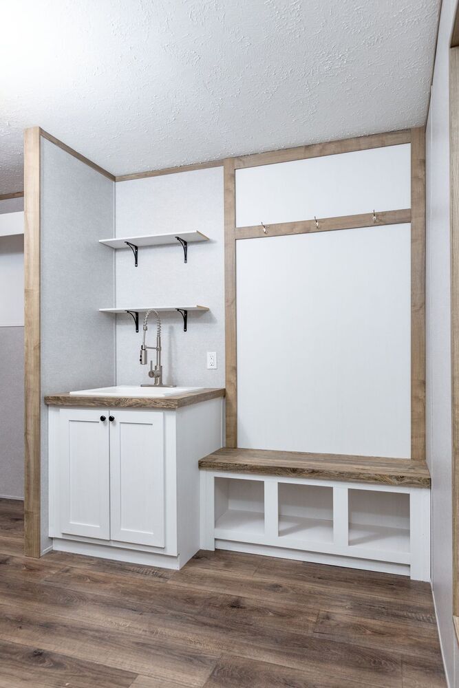 Laundry room setup with a white cabinet and sink, wooden bench with storage, coat hooks above, and two shelves on the wall. Neutral, modern style.