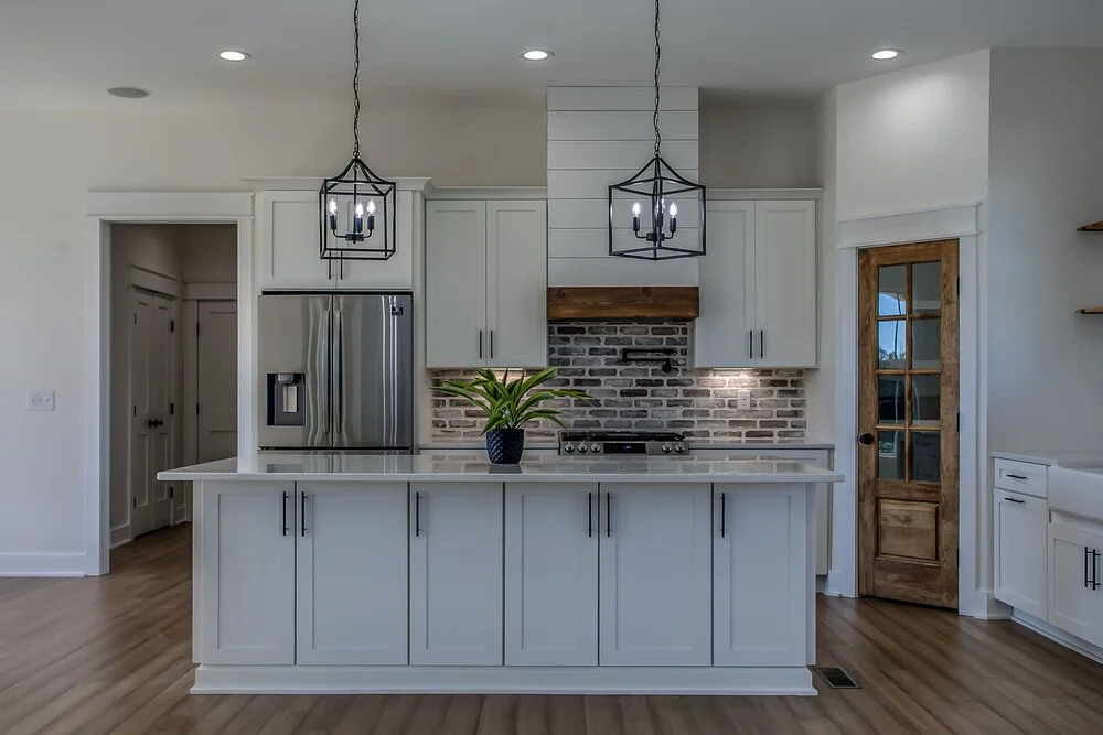 Modern kitchen with white cabinets, stainless steel fridge, and brick backsplash. A wooden door adds warmth. Two black pendant lights hang above a central island with a green plant.