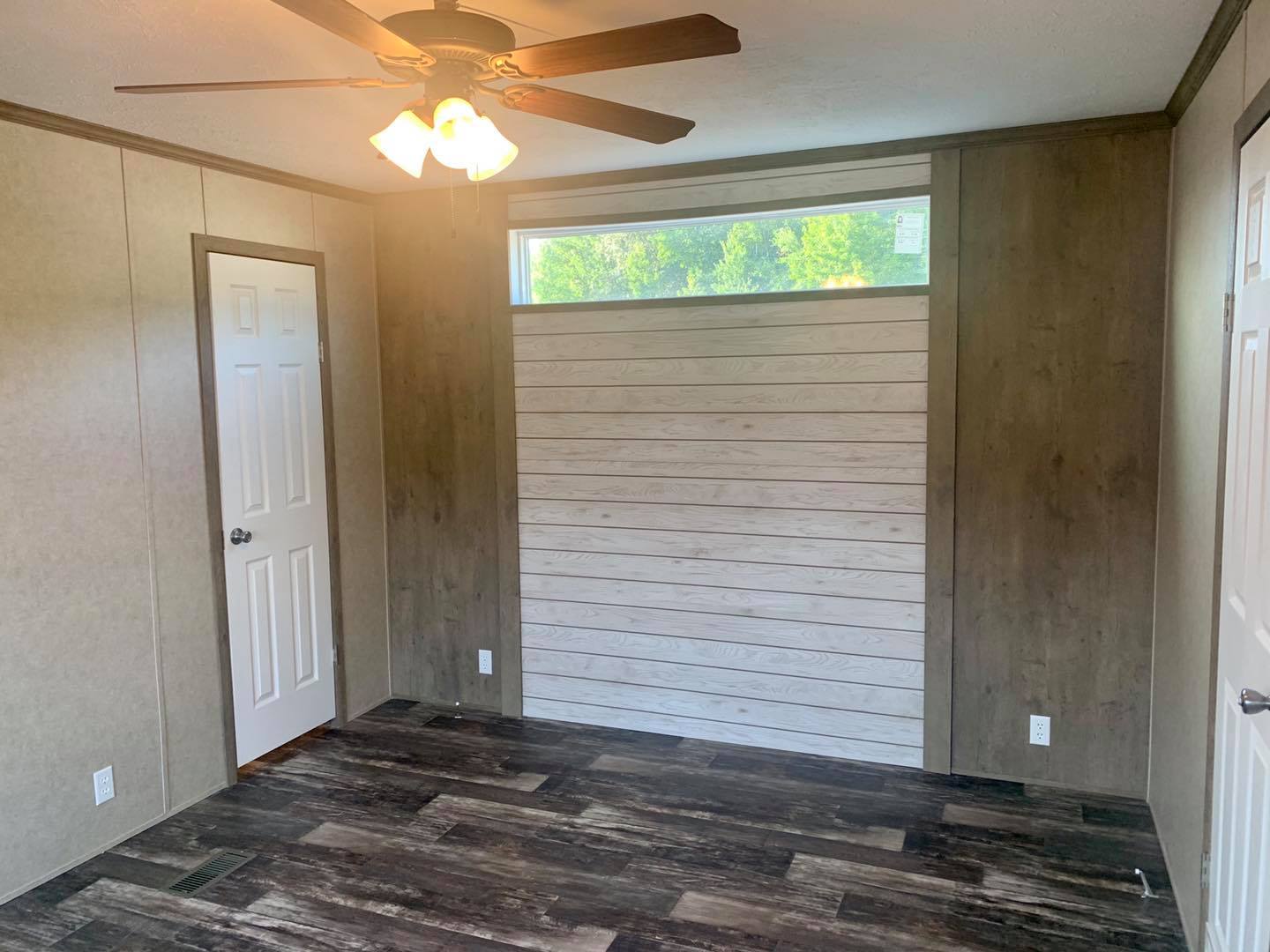 Empty room with a ceiling fan, wood panel walls, and wood-plank flooring. Two doors and a rectangular window offer natural light, creating a calm atmosphere.