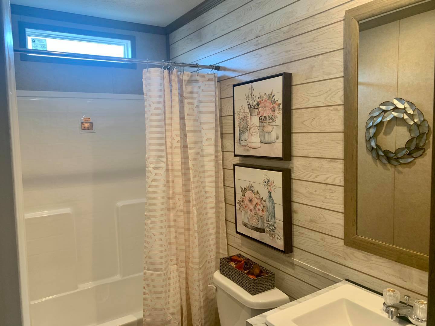 Cozy bathroom with a striped shower curtain, wooden panel walls, and floral artwork. A decorative wreath mirror and a basket add rustic charm.