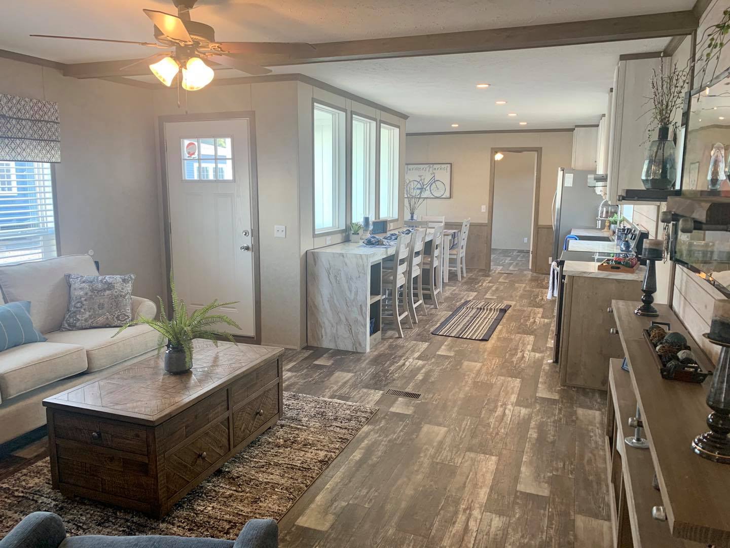 A cozy living room with a beige sofa and blue cushions. A rustic wooden coffee table and a potted plant are in front. Bar stools line a counter leading to the kitchen. Neutral tones and warm lighting create a welcoming atmosphere.