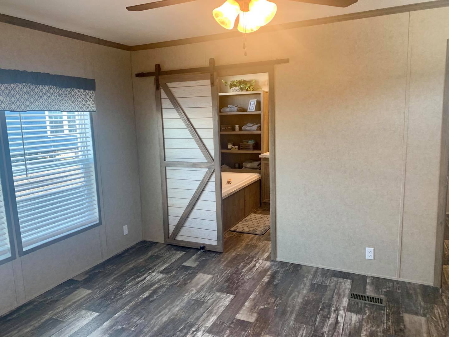 Room with wood-look flooring, a ceiling fan with a light, and a large window with a patterned valance. A sliding barn door reveals a storage area. The tone is cozy and rustic.