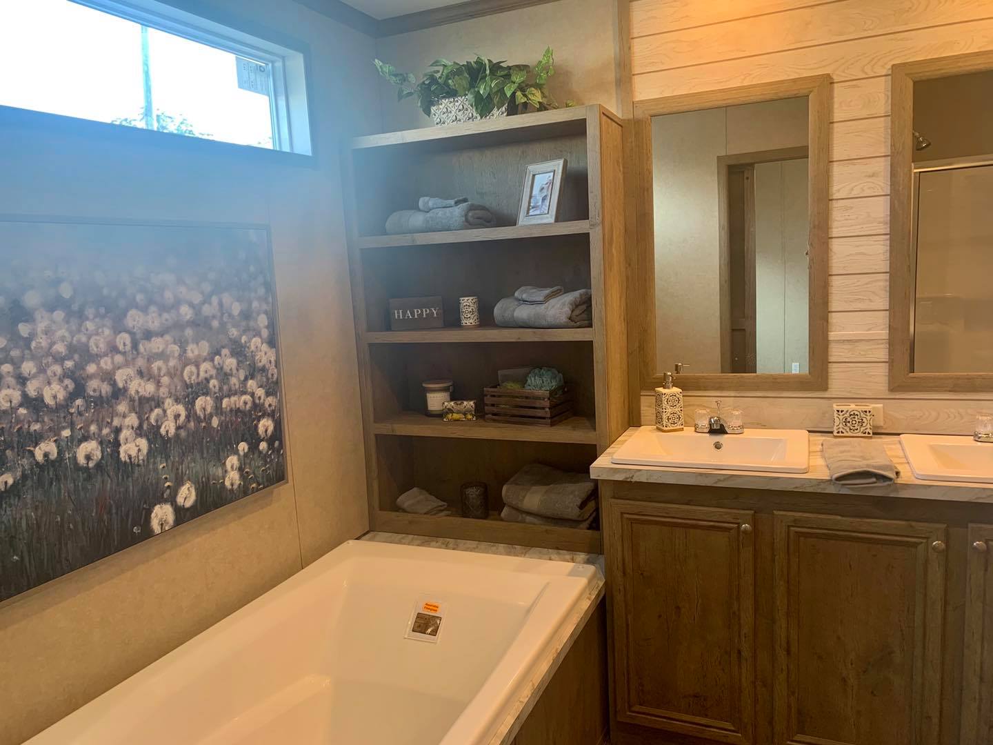 Cozy bathroom with a white tub and dual sinks. Shelves hold towels, plants, and decor. A serene flower painting adds elegance. Natural light.