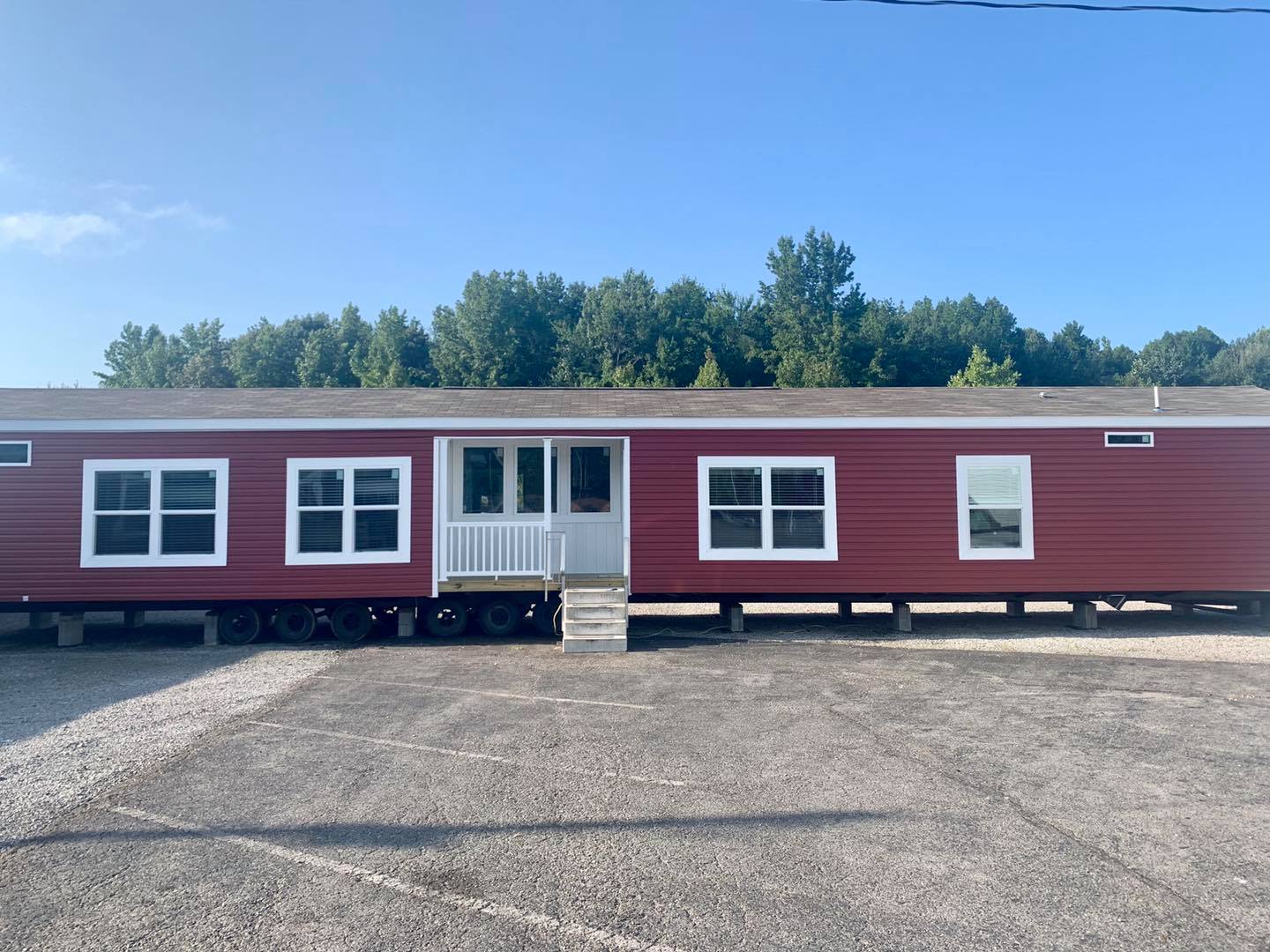 A red mobile home with multiple windows is set on a gravel lot. There are stairs leading to the entrance. The background features green trees under a clear blue sky.