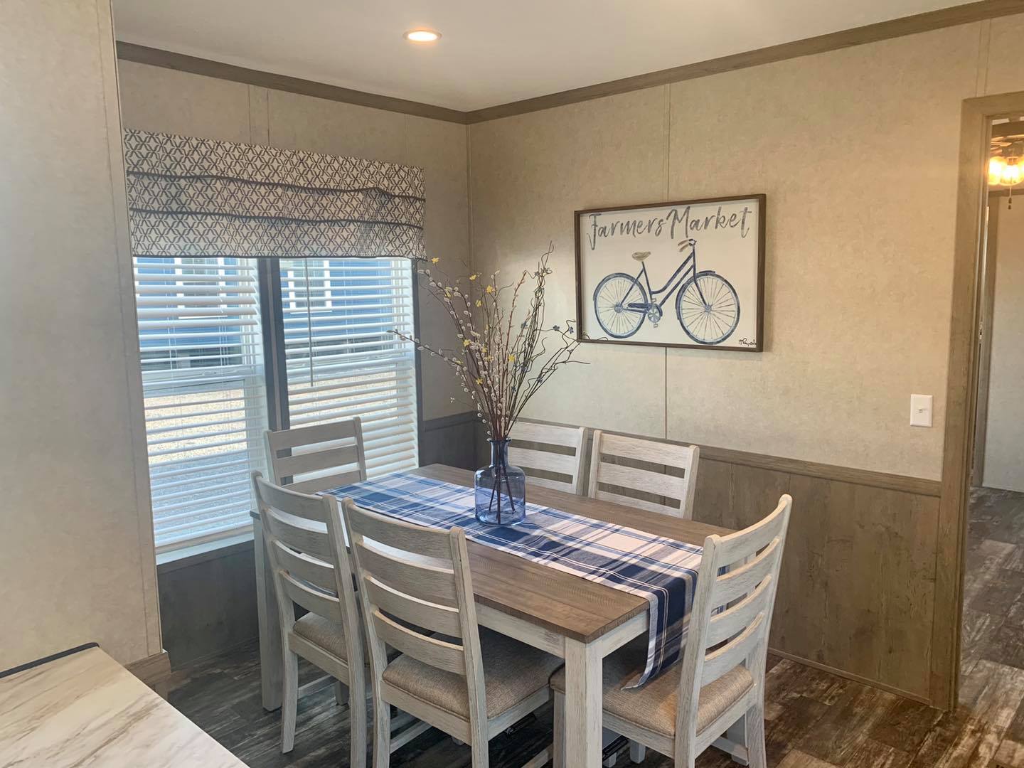 Dining area with a wooden table, six chairs, and a blue plaid runner. A vase with branches is on the table. A "Farmer's Market" bike sign hangs on the wall. Cozy ambiance.