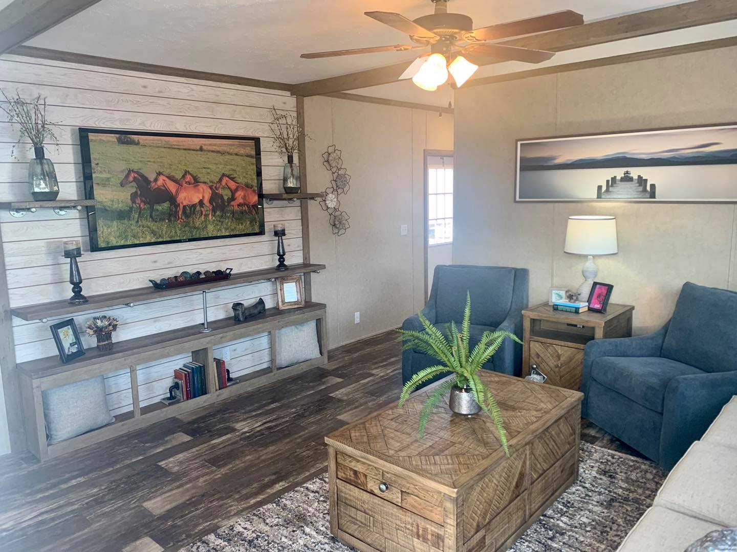 Cozy living room with wooden floors and beige walls. Features blue chairs, wooden furniture, horse painting, landscape photo, and modern decor. Warm lighting.