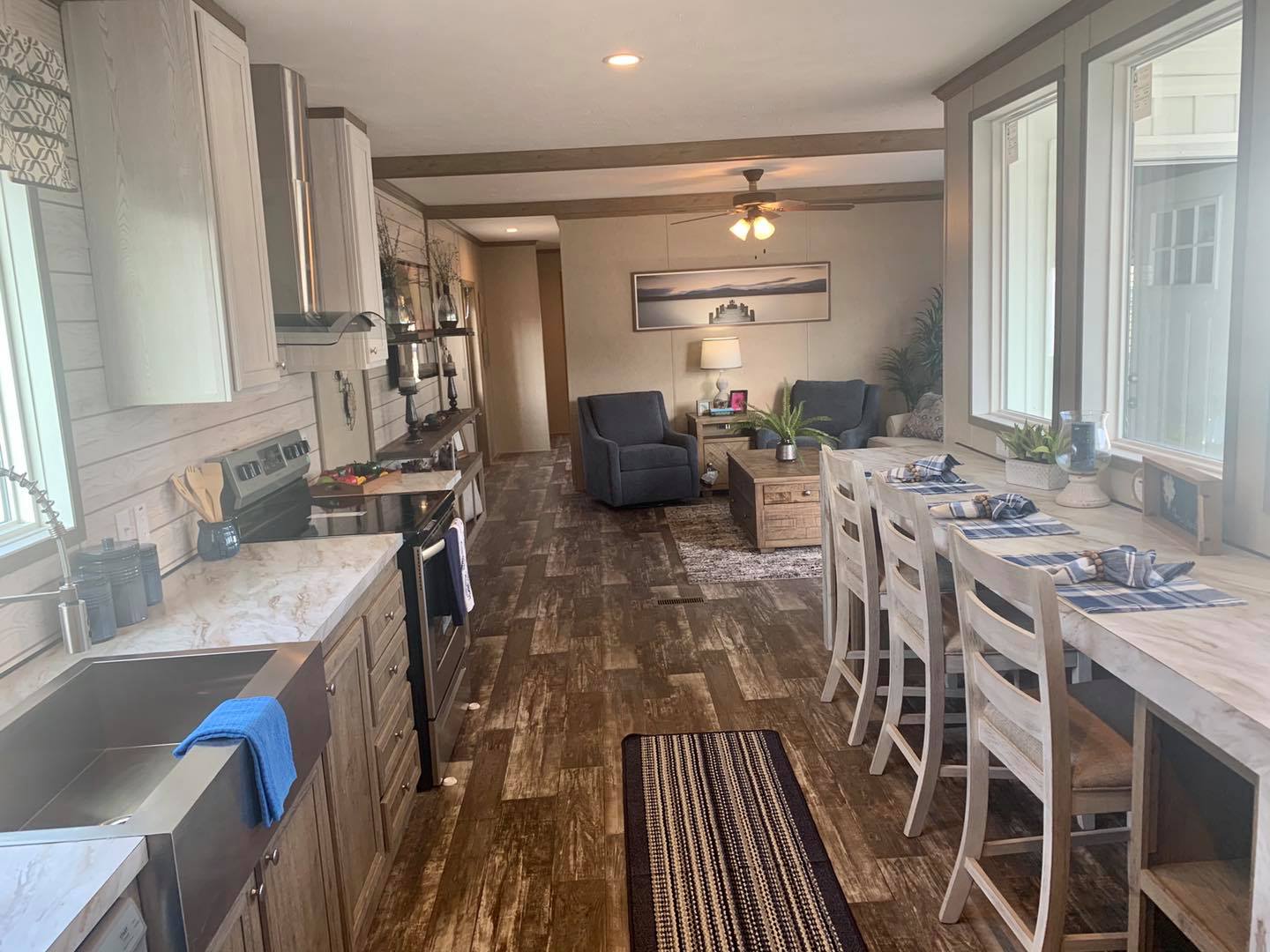 Warmly lit open kitchen and living area. Rustic wood floors, marble countertops, breakfast bar with stools, and cozy seating by a large window. Relaxed ambiance.