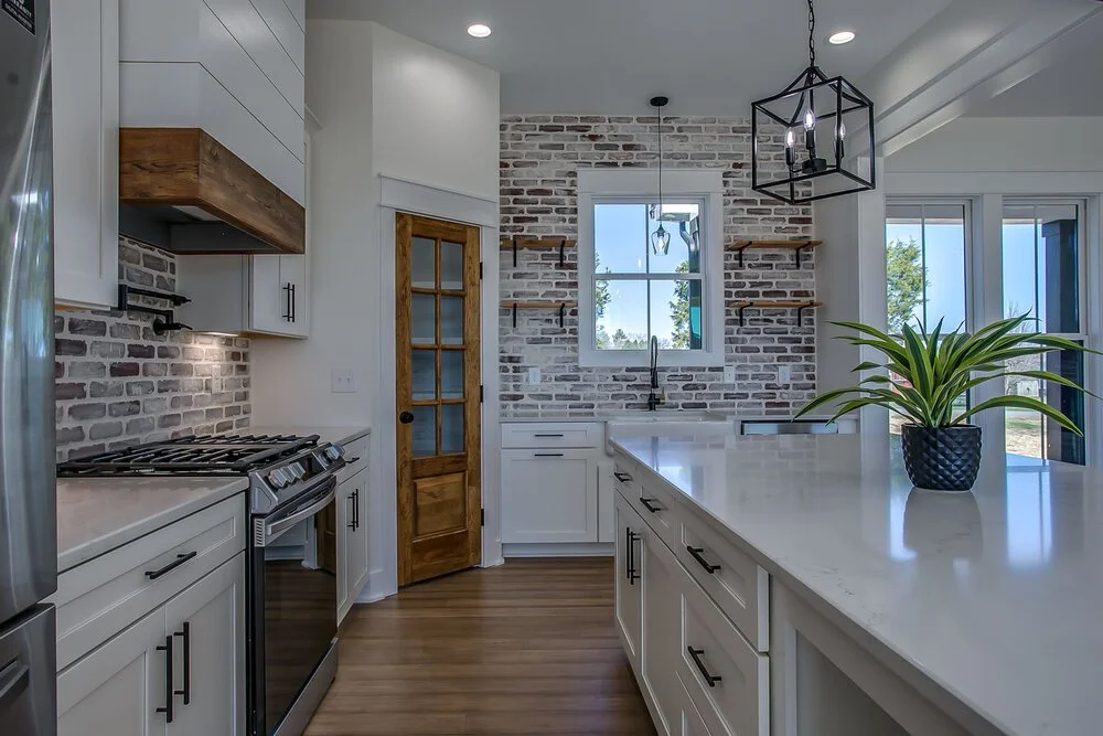 Modern kitchen with white cabinets, light wood flooring, and brick backsplash. A potted plant on the island adds freshness, and a large window brightens the space.