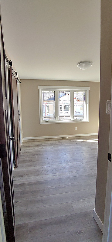 Bright, empty room with light wooden floors and beige walls. A large window on the far wall lets in sunlight, creating a warm, welcoming atmosphere.