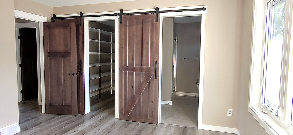 Room with light wood floors, beige walls, two open sliding barn doors revealing a closet with shelves and a bathroom. Large window on the right.
