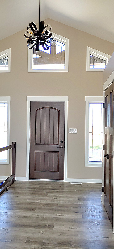 Bright entryway featuring a wooden door framed by tall windows. A modern black chandelier hangs from a high ceiling, casting soft light on hardwood floors.
