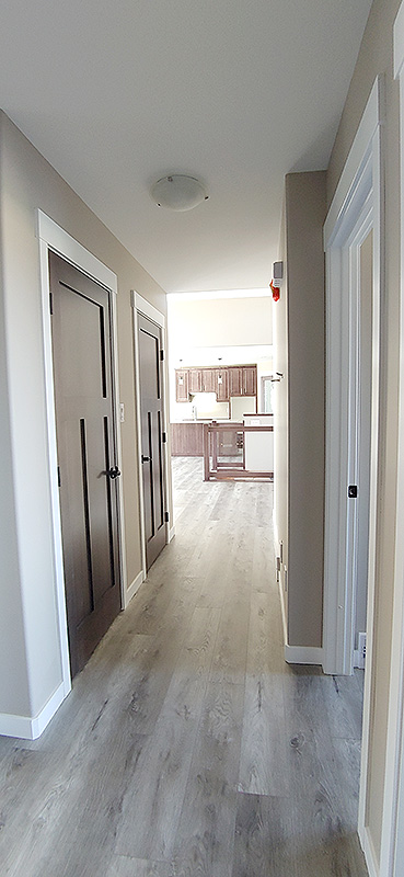 A well-lit hallway with light wood flooring leads to a kitchen. The walls are beige, and dark wooden doors are on the left. The kitchen features wooden cabinets.