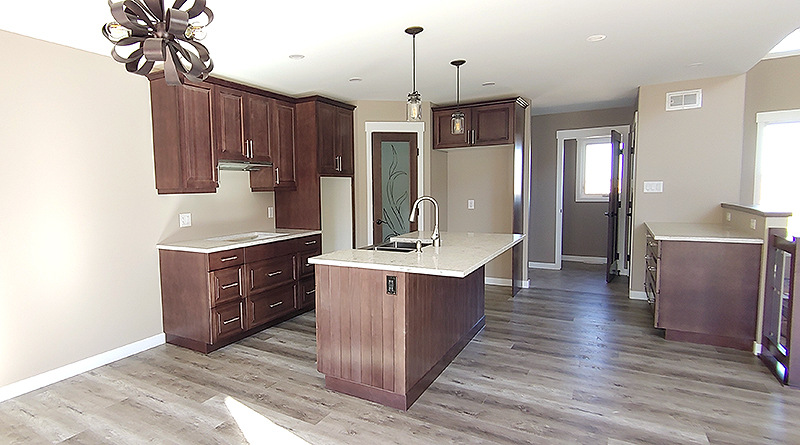 Modern kitchen with dark wood cabinets, marble countertops, and a center island. Features pendant lighting, wood flooring, and beige walls. Bright, spacious ambiance.