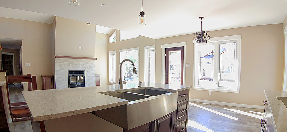 Spacious kitchen with beige walls, center island featuring a large stainless steel sink, pendant lights, and ample natural light through large windows. Cozy and inviting.