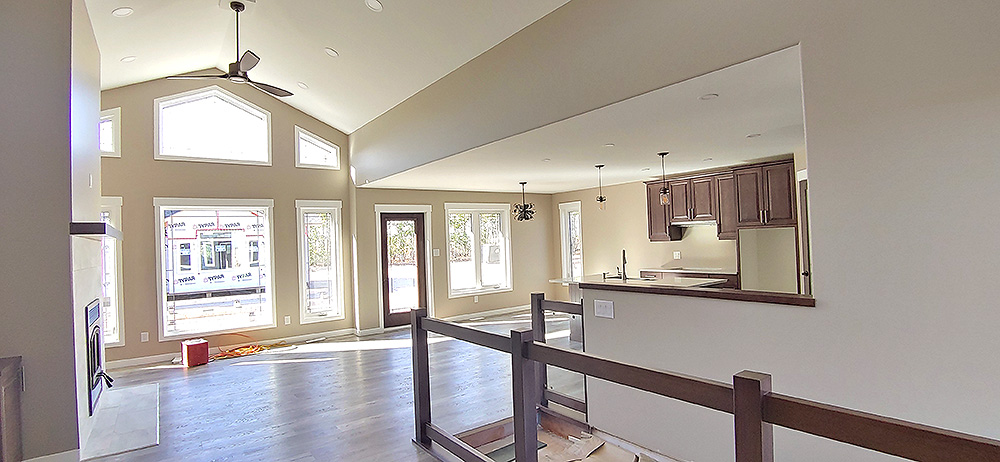 Spacious, bright living area with large windows, vaulted ceiling, and ceiling fan. Kitchen with wooden cabinets visible. Modern, welcoming atmosphere.