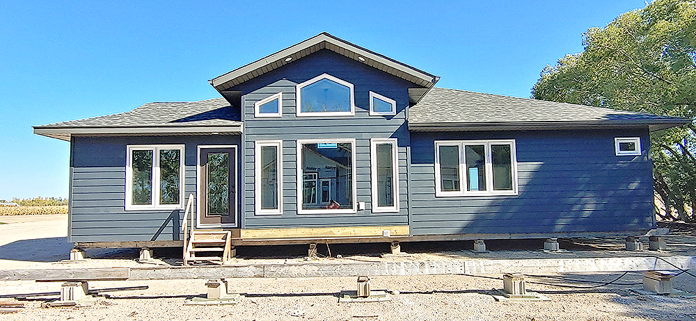 Blue, modern prefab home on pilings, with large windows and a gabled roof, set against a clear blue sky and trees to the right, evoking a serene atmosphere.