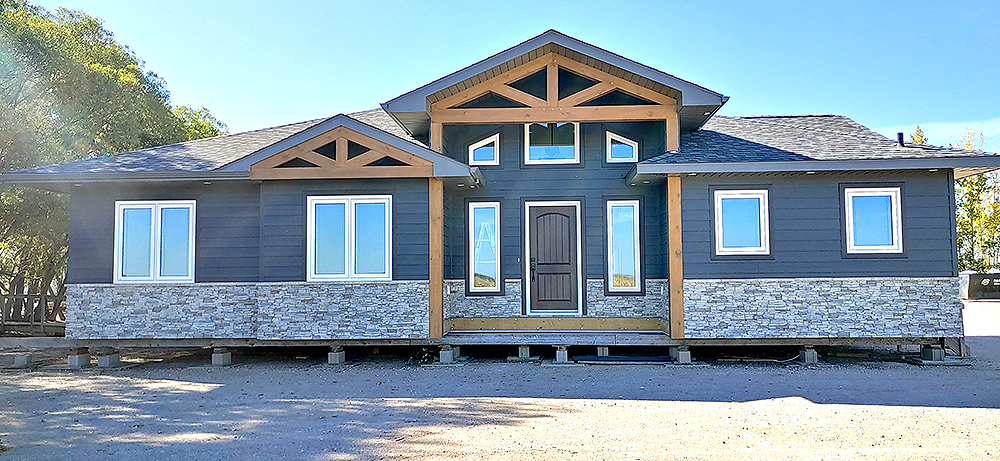 Single-story prefab home with charcoal siding, stone accents, peaked roof, white-framed windows, and landscaped surroundings under a clear blue sky.