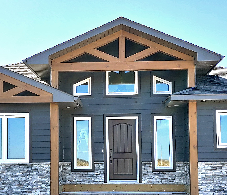 Front of a modern house with gray siding and stone accents. A dark wooden door is centered beneath a gabled roof. Bright, sunny day.