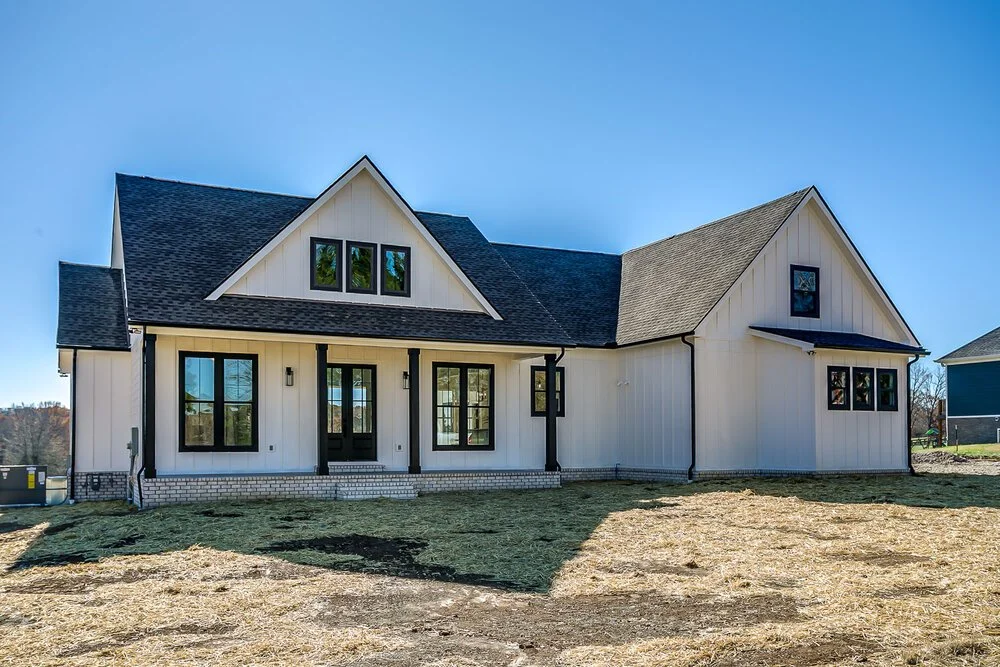 A modern farmhouse with white siding and black trim, featuring a gabled roof and front porch. The yard is bare, suggesting recent construction. Bright, sunny day.